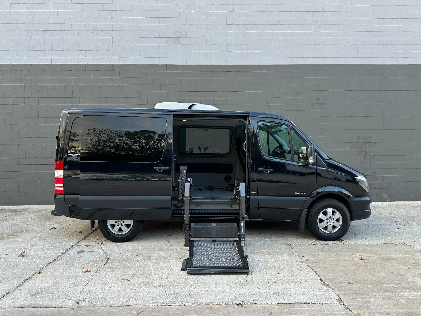 Black Mercedes Sprinter full size wheelchair van with lift deployed from open passenger side sliding door.