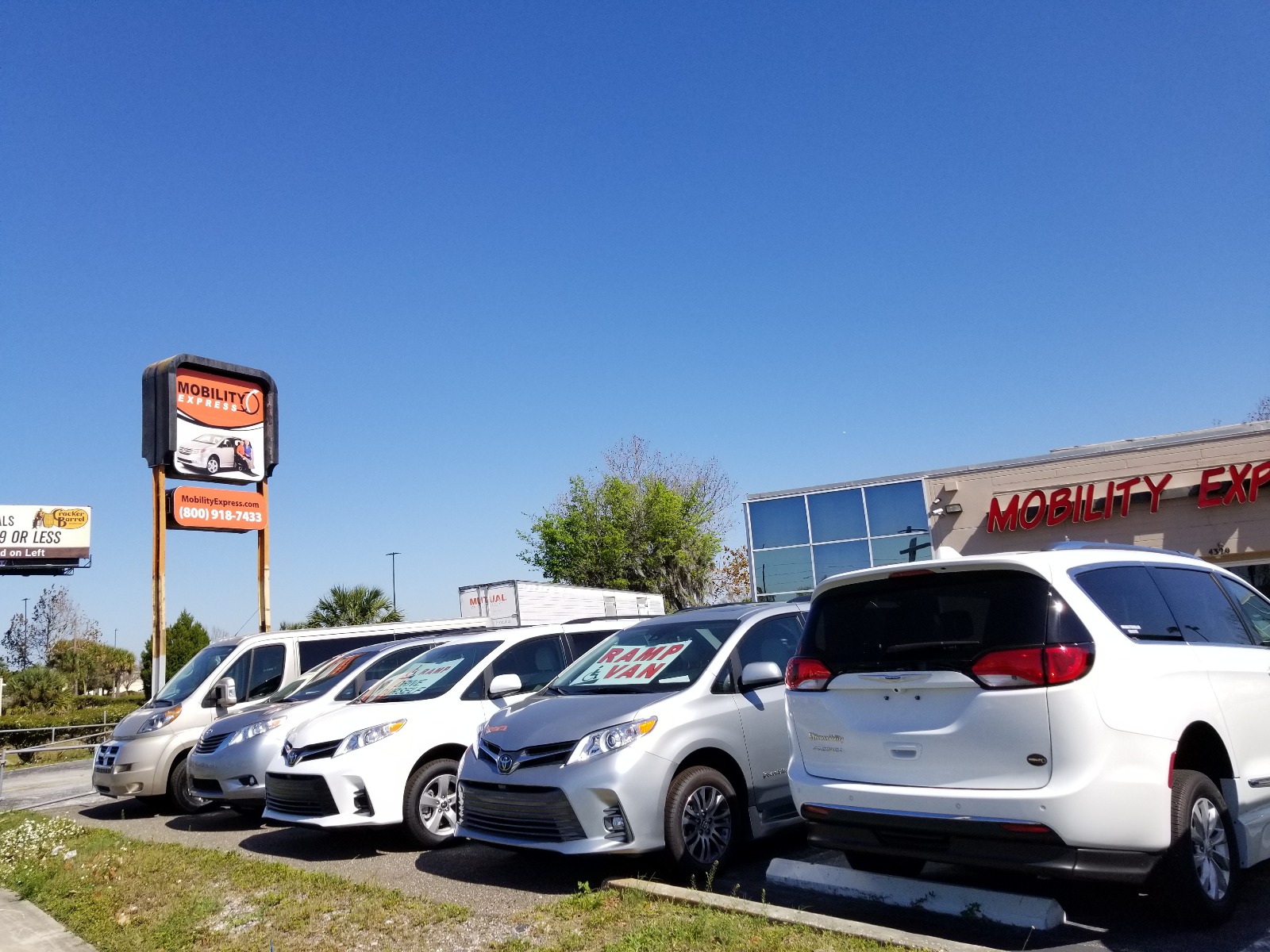 A row of wheelchair vans, most facing forward, under a cloudless blue sky. There's an orange sign in the background to the left that says Mobility Express, along with the website and phone number.