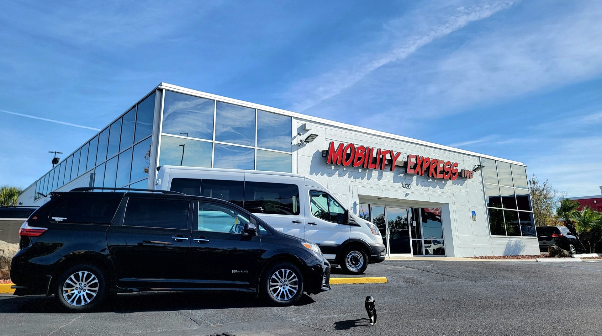 A white building with a lot of mirrored windows with the words "Mobility Express" in red on the front, under a blue sky with light clouds. There are two vans parked in front of the building to the left, a dark colored minivan and a white full size van.