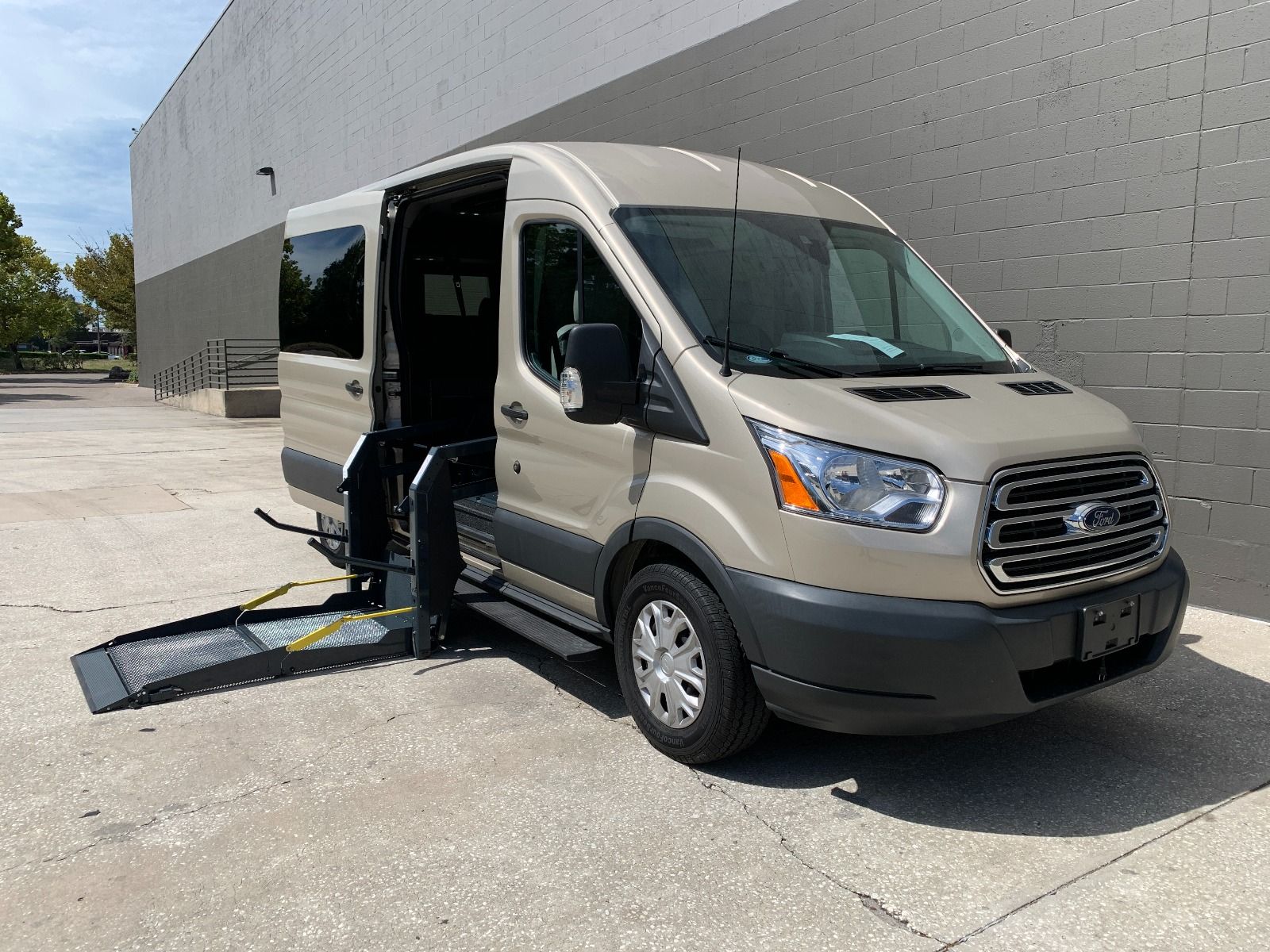 Passenger side of a gold 2017 Ford Transit Full Size Wheelchair Van, with lift deployed, as seen from front angle