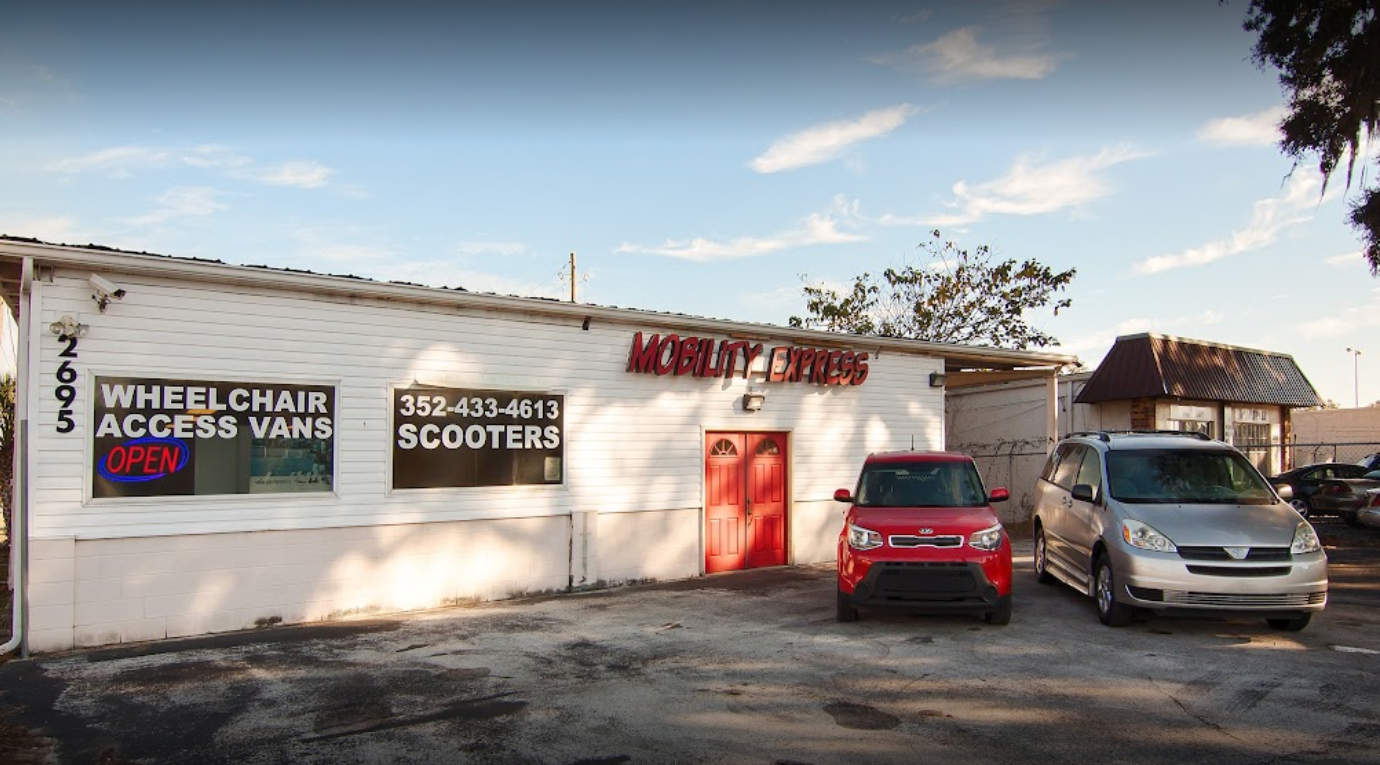 A white building with the words "Mobility Express" across the top. There are two wheelchair accessible vehicles parked to the right, a red small handicap accessible SUV, and a silver wheelchair accessible minivan.