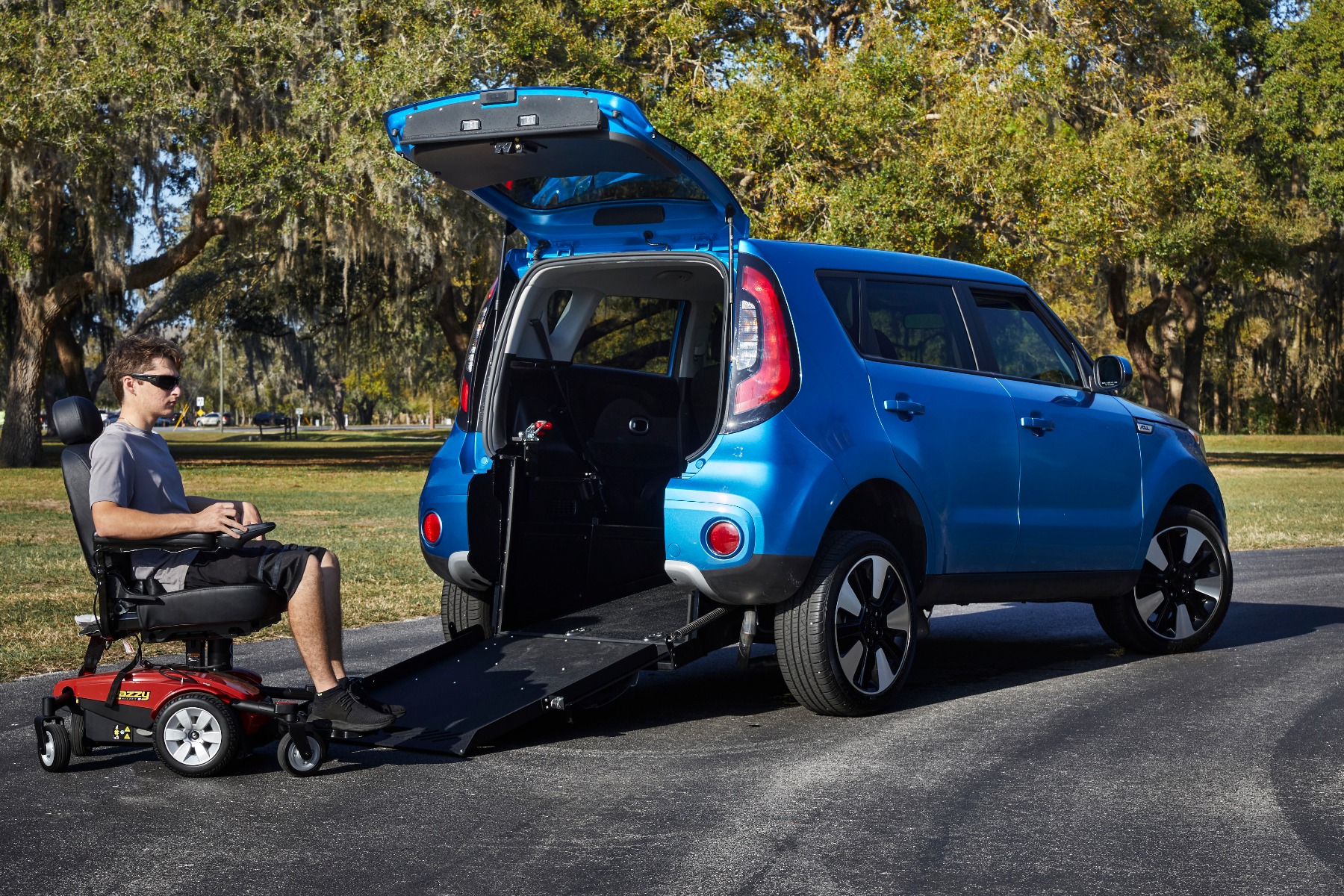 A man is sitting on a red power wheelchair. The power wheelchair is at the bottom edge of a ramp deployed from the rear hatch of a wheelchair accessible Kia Soul small SUV. The Kia Soul is blue. The setting seems to be a park with trees and a road in the background.