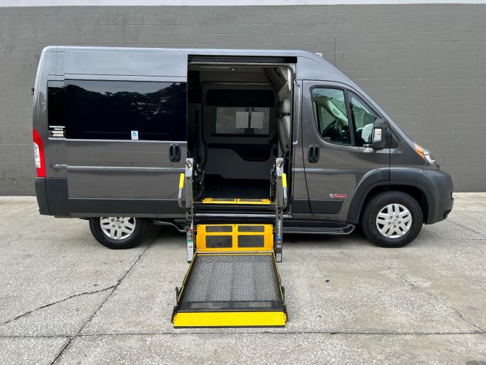 Gray RAM ProMaster full size wheelchair van with lift deployed from open rear barn doors. Large gray wall in the background.