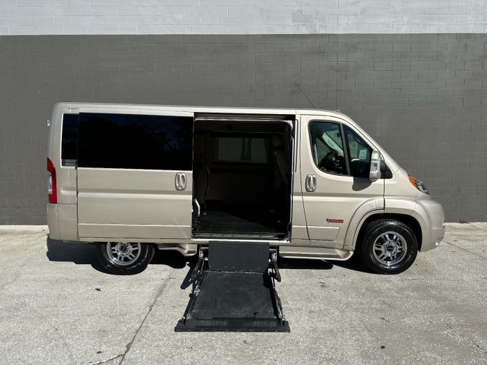 Gold RAM ProMaster full size wheelchair van with lift deployed from open rear barn doors. Large gray wall in the background.