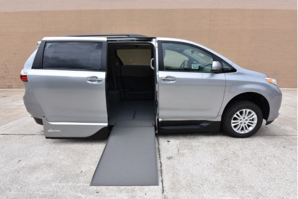A Toyota Sienna Wheelchair Van in Silver, seen from the passenger side, with the wheelchair ramp deployed from the open passenger side sliding door.
